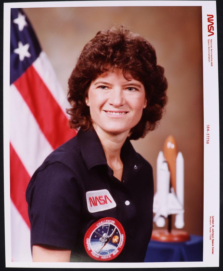 Color photo portrait of a woman with chin length curly brown hair, wearing a navy blue short sleeved polo shirt with two patches. Top patch: NASA name logo.  Bottom patch: Circular mission patch with Challenger Space shuttle and astronaut's name.  Blurred Background: on the left American flag, on the ride Space Shuttle model. Border: NASA photo markings.
