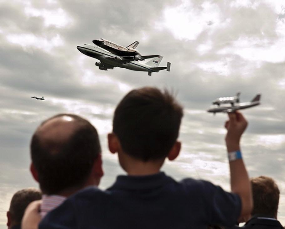 Back view of a child and adult watching a plane with the Space Shuttle Discovery attached fly over the Washington, D.C. area.